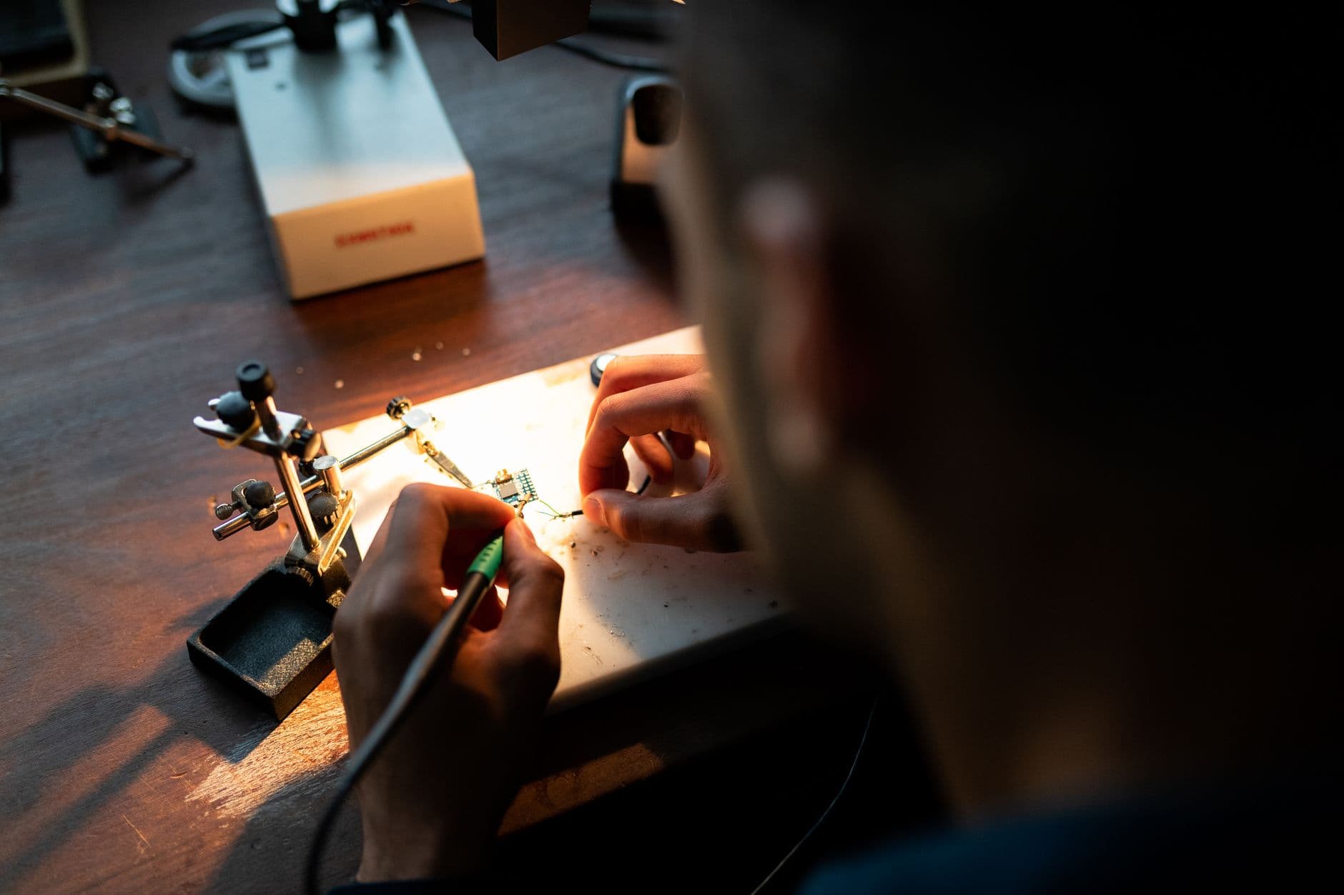 A maker working with a soldering iron and helping hands at a lamp-lit workbench