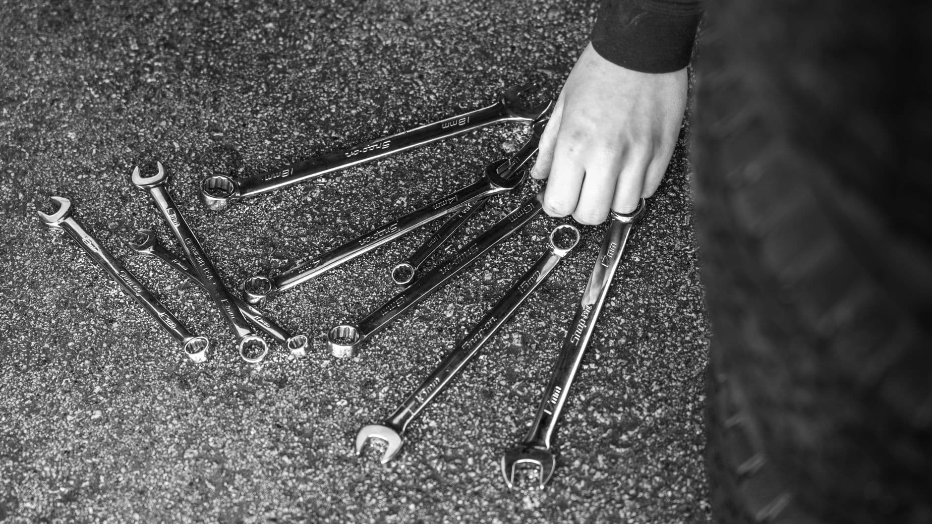 A hand fanning out a set of combination wrenches on rough asphalt, in black and white