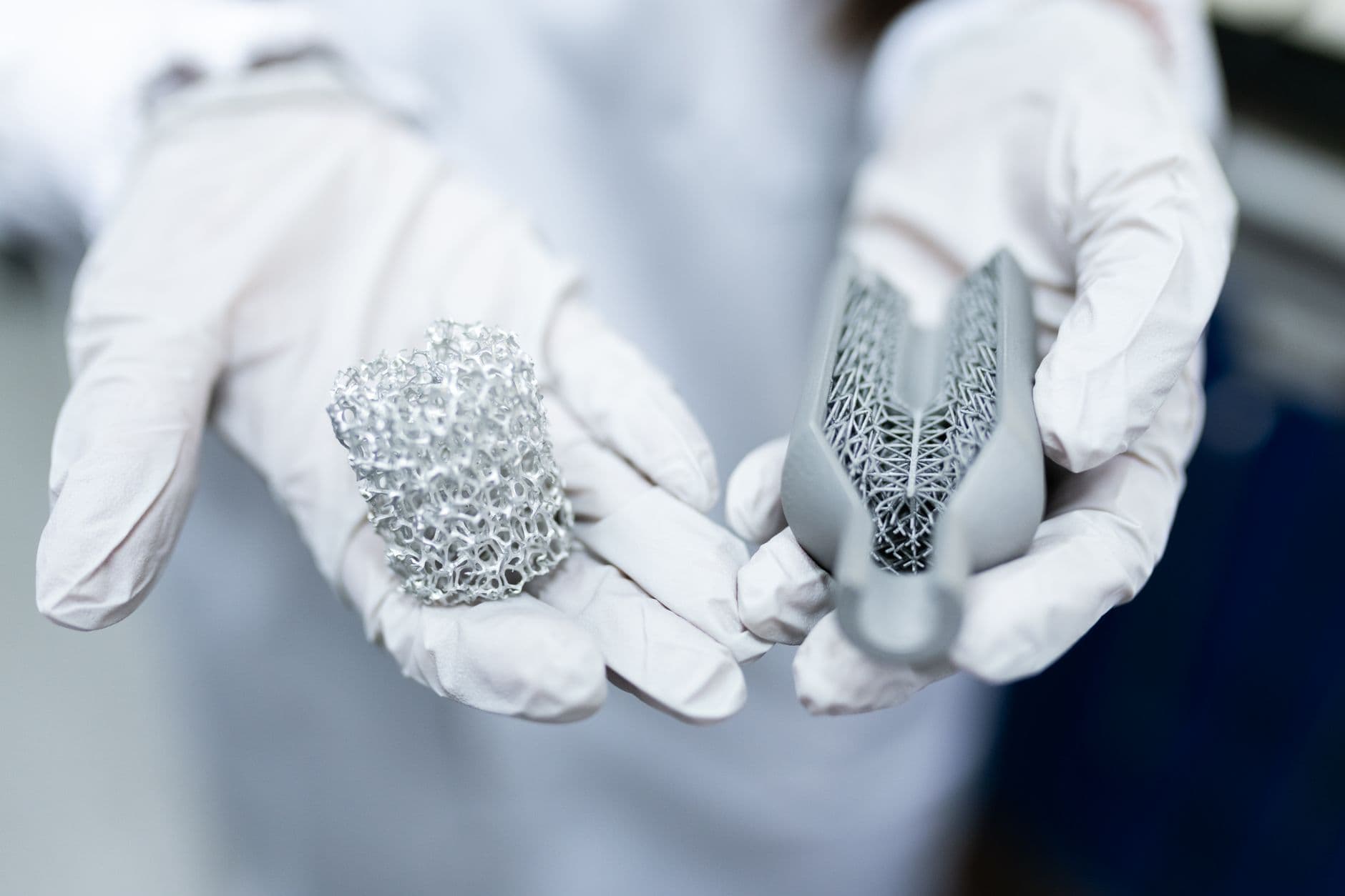 Gloved hands holding two 3D-printed metal lattice parts in a lab